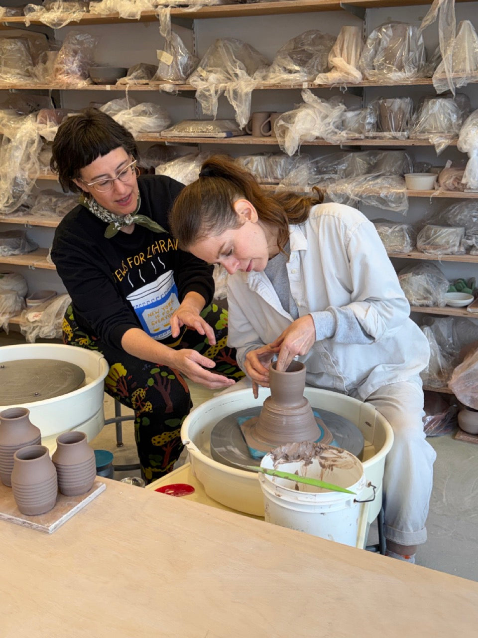 Two women in a pottery studio, one shaping a clay vessel on a pottery wheel while the other observes and guides. Shelves in the background are filled with plastic-wrapped pottery pieces.