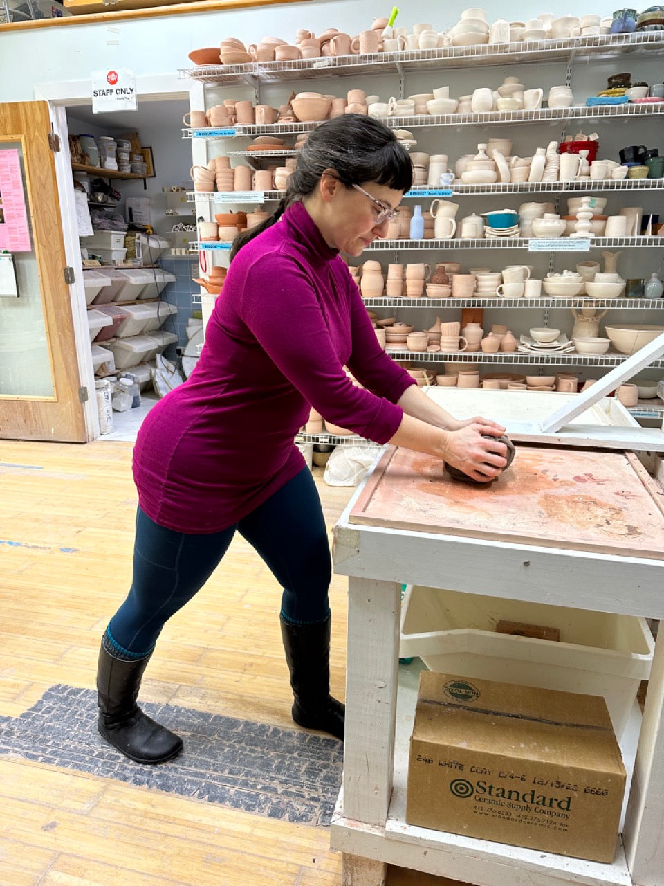 A woman in a purple long-sleeve top, dark pants, and black boots is wedging clay on a wooden table inside a pottery studio. Shelves in the background are filled with ceramic pottery pieces.